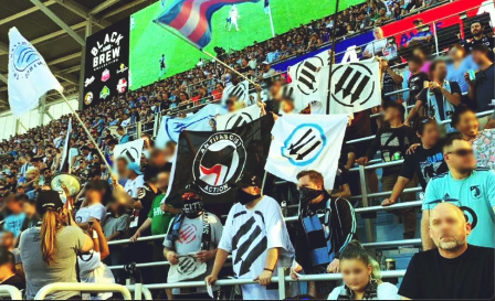 Photo of Portland Timbers fans holding flags depicting three arrows and written description.