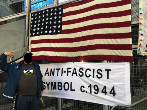 Photo of soccer fan with 48-star American flag and Iron Front shirt, and sign stating: Anti-fascist symbol circa 1944.
